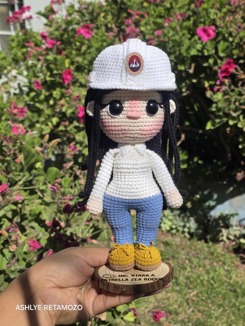 May include: A crochet doll featuring long black hair, a white hard hat with a small emblem, a white long-sleeved shirt, blue pants, and yellow shoes. The doll stands on a circular wooden base with the text ING. KIARA A. ESTRELLA ZEA ROQUE.