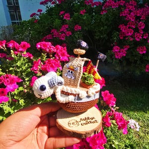 Puede incluir: Un barco de ganchillo con un casco blanco y marrón, una vela beige con una calavera y huesos cruzados, y una figura de proa de animal blanco. El barco está sobre una base de madera con el texto "Ashlye crochet".