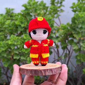 Pode incluir: Uma boneca de bombeiro de crochê vestindo um chapéu vermelho com o número 18. A boneca está vestindo um uniforme de crochê vermelho e amarelo e está em pé sobre uma base de madeira.