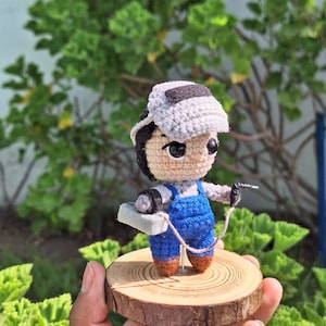 Puede incluir: Figura amigurumi de crochet de un soldador con un mono azul, una máscara de soldar y una antorcha de soldar. La figura está de pie sobre una base de madera.