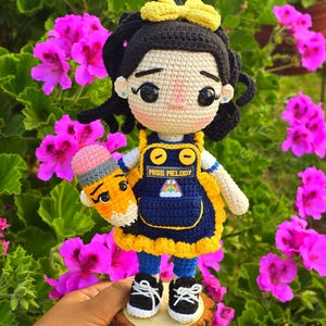 Puede incluir: Una muñeca de ganchillo con pelo negro, un lazo amarillo y tez beige. La muñeca lleva un vestido de peto azul con ribete amarillo, con el texto MISS MELODY y un parche de arcoíris. Sostiene un lápiz de ganchillo y lleva zapatillas negras.