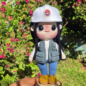 May include: A crochet doll featuring a white hard hat with a circular emblem, black hair, a white shirt, a gray utility vest, blue pants, and mustard yellow shoes. The doll stands on a circular wooden base engraved with the text ING. KIARA A. ESTRELLA ZEA ROQUE.
