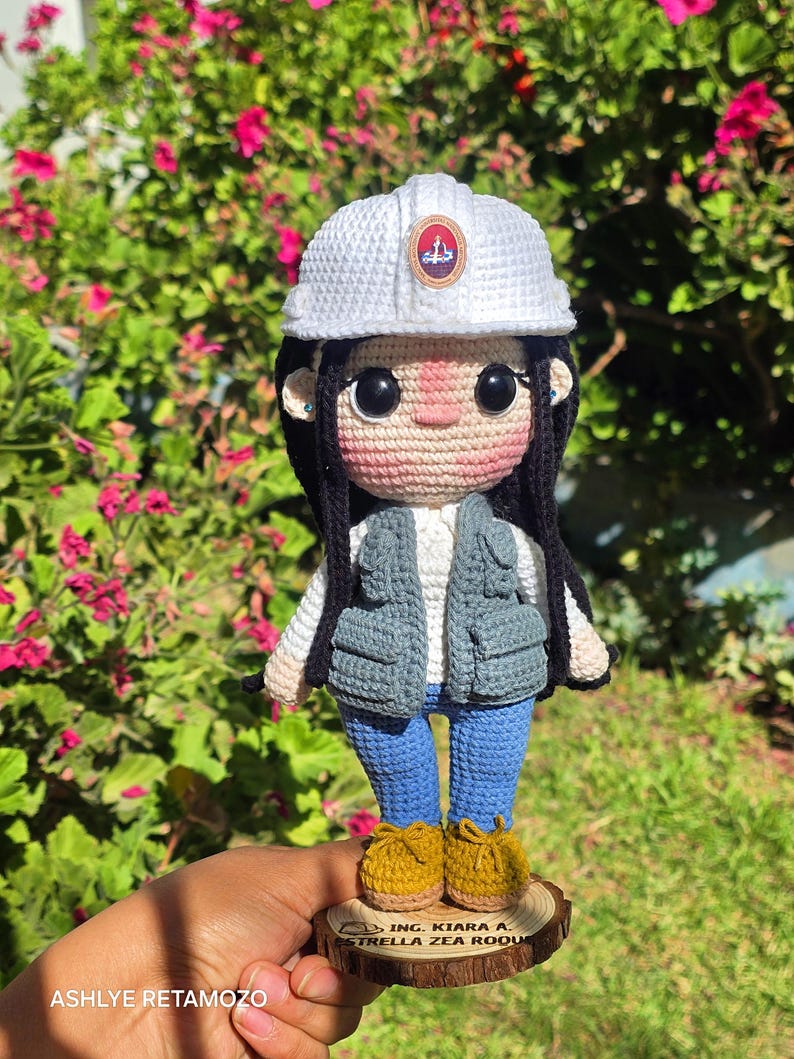 May include: A crochet doll featuring a white hard hat with a circular emblem, black hair, a white shirt, a gray utility vest, blue pants, and mustard yellow shoes. The doll stands on a circular wooden base engraved with the text ING. KIARA A. ESTRELLA ZEA ROQUE.