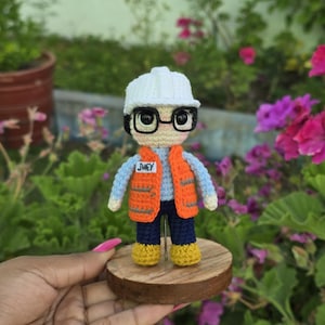 Op de afbeelding: Een gehaakte pop met een witte helm, een blauw shirt, een oranje vest, een blauwe broek en gele schoenen. De pop houdt een naamsticker vast met de tekst "Jhey".