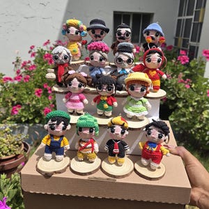 May include: A collection of crocheted dolls, each with unique outfits and hairstyles, displayed on wooden platforms. The dolls are arranged on a cardboard box, with a background of pink flowers and greenery. The dolls are of various sizes and colors.