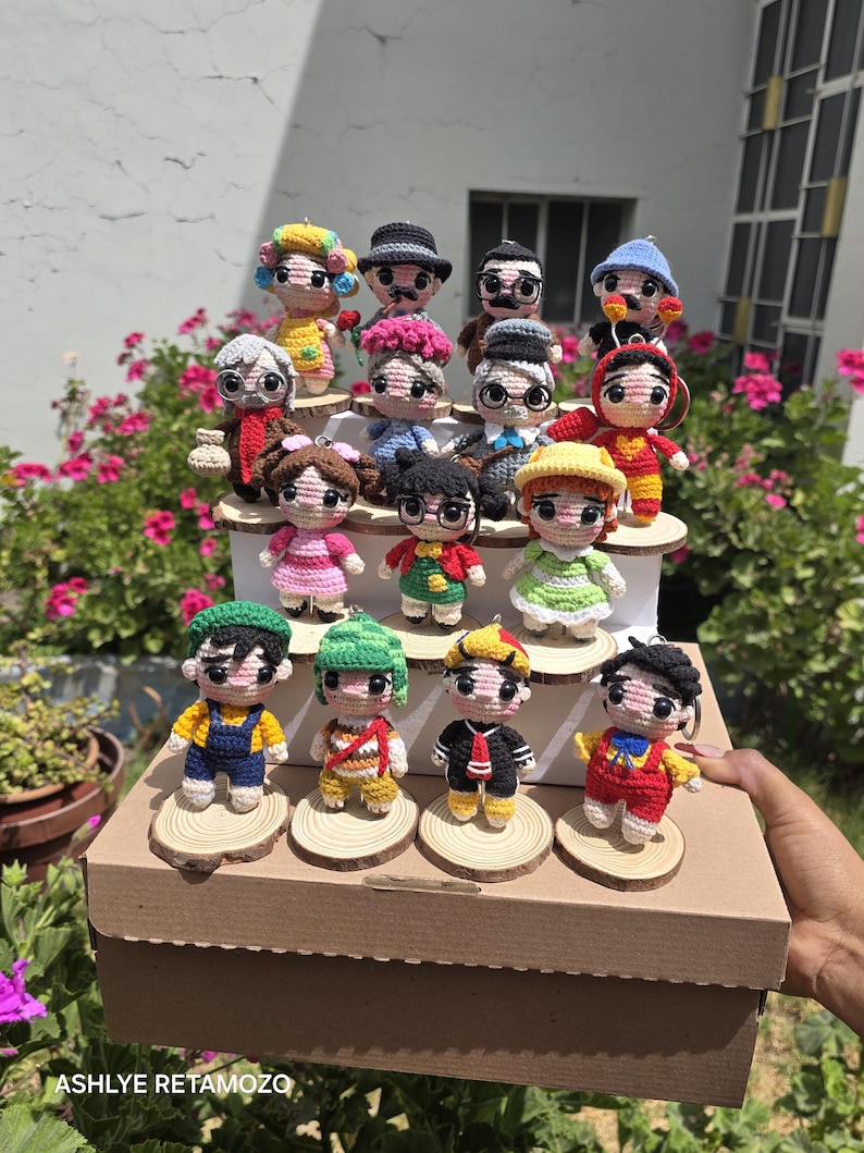May include: A collection of crocheted dolls, each with unique outfits and hairstyles, displayed on wooden platforms. The dolls are arranged on a cardboard box, with a background of pink flowers and greenery. The dolls are of various sizes and colors.