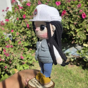 May include: A crochet doll featuring long black hair, a white hard hat with a circular logo, a gray vest, a white long-sleeved shirt, blue pants, and yellow shoes. The doll stands on a circular wooden base with the text ING. KIARA A. ESTRELLA ZEA ROQUE.