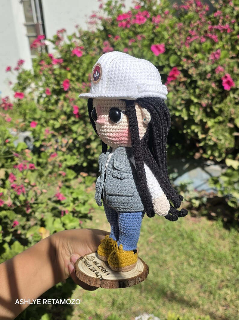 May include: A crochet doll featuring long black hair, a white hard hat with a circular logo, a gray vest, a white long-sleeved shirt, blue pants, and yellow shoes. The doll stands on a circular wooden base with the text ING. KIARA A. ESTRELLA ZEA ROQUE.