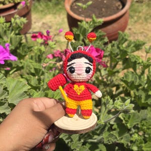 May include: A handmade crochet doll in red and yellow, holding a yellow hammer. The doll has a heart on its chest and is mounted on a small wooden disc. The doll has a red headpiece with yellow accents and two antennae.