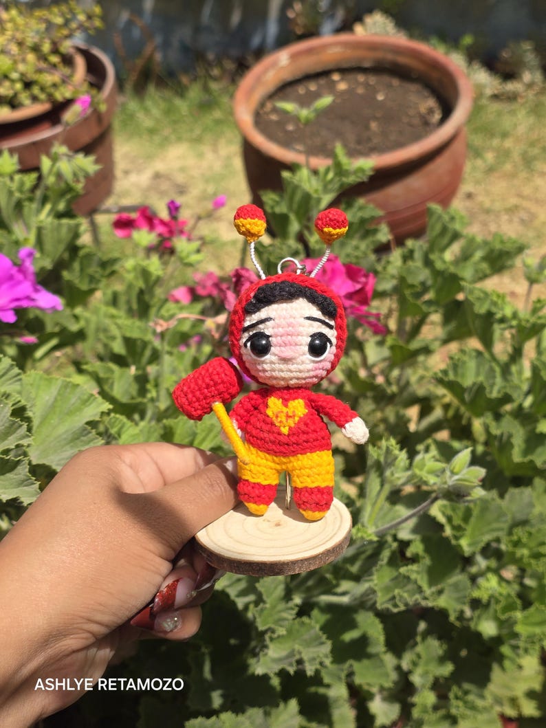 May include: A handmade crochet doll in red and yellow, holding a yellow hammer. The doll has a heart on its chest and is mounted on a small wooden disc. The doll has a red headpiece with yellow accents and two antennae.
