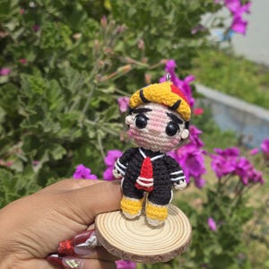 May include: A handmade crochet doll of a cartoon character, wearing a black sailor suit with white stripes, a red tie, and yellow shoes. The doll has a yellow and red hat and is mounted on a small wooden disc. The background features green foliage and purple flowers.