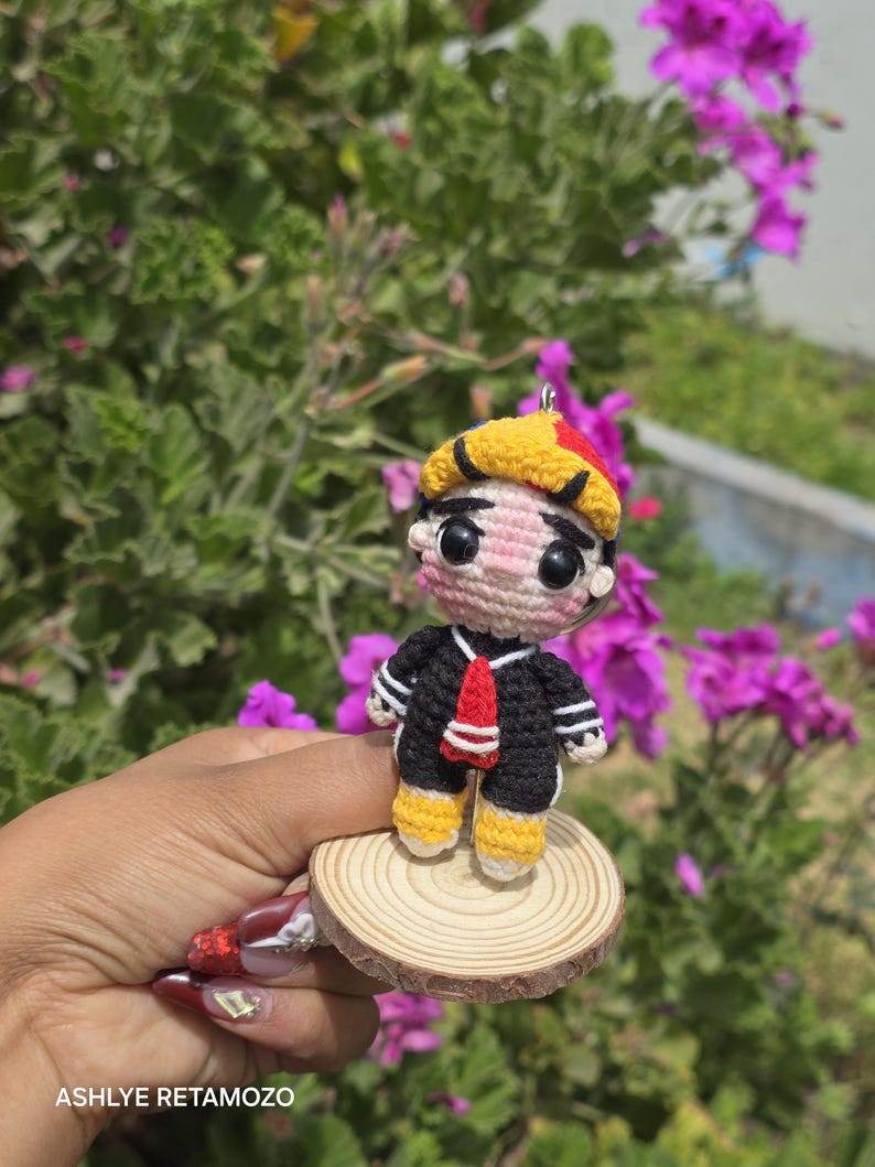 May include: A handmade crochet doll of a cartoon character, wearing a black sailor suit with white stripes, a red tie, and yellow shoes. The doll has a yellow and red hat and is mounted on a small wooden disc. The background features green foliage and purple flowers.