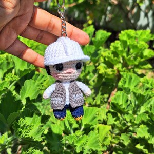 May include: A crochet keyring featuring a small figure wearing a white hard hat, a grey waistcoat over a white shirt, and dark blue trousers. The figure has large black eyes and is attached to a silver metal keyring with a short chain link connector.