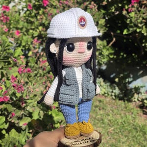 May include: A crochet doll featuring long black hair, a white hard hat with a circular emblem, a grey utility vest, a white shirt, blue pants, and yellow shoes. The doll stands on a circular wooden base engraved with the text ING. KIARA A. ESTRELLA ZEA ROQUE.