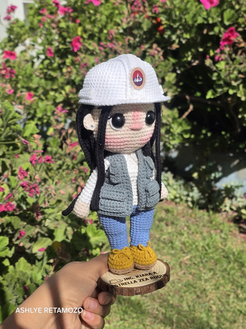 May include: A crochet doll featuring long black hair, a white hard hat with a circular emblem, a grey utility vest, a white shirt, blue pants, and yellow shoes. The doll stands on a circular wooden base engraved with the text ING. KIARA A. ESTRELLA ZEA ROQUE.