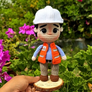 May include: A crochet doll of a construction worker wearing a white hard hat, a bright orange safety vest with reflective silver stripes, a light blue long-sleeved shirt, brown trousers, and brown shoes, standing on a circular wooden base.