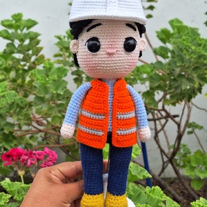 May include: A crocheted doll wearing a white hard hat, blue shirt, orange safety vest, blue pants, and yellow shoes. The doll is standing on a white base.