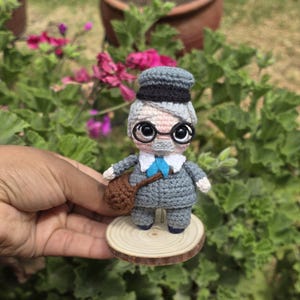 May include: A handmade crochet doll of an elderly gentleman, wearing a gray suit, a blue bow tie, and a gray and black hat. The doll has large, round eyes and wire-rimmed glasses. It is holding a brown satchel and stands on a small wooden disc.