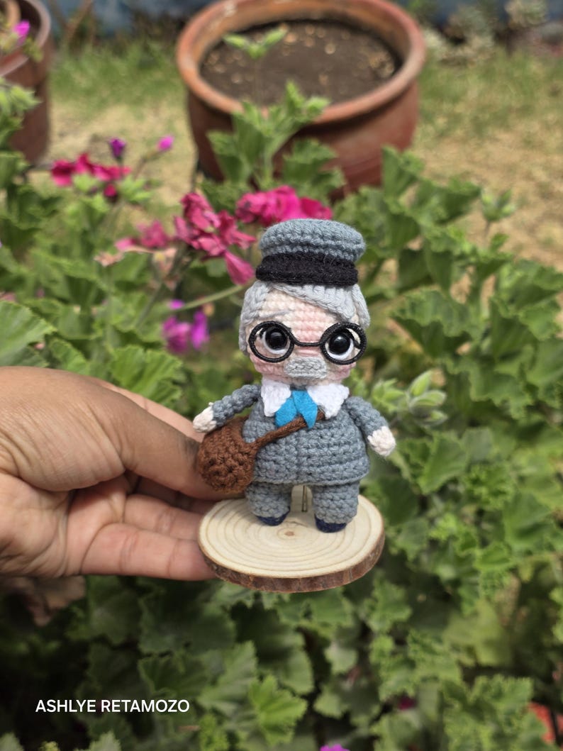 May include: A handmade crochet doll of an elderly gentleman, wearing a gray suit, a blue bow tie, and a gray and black hat. The doll has large, round eyes and wire-rimmed glasses. It is holding a brown satchel and stands on a small wooden disc.