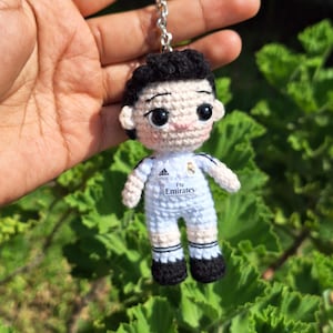 Puede incluir: Llavero de ganchillo de un futbolista que lleva una camiseta blanca con detalles negros y el texto "Fly Emirates".