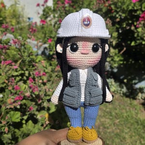May include: A crocheted doll wearing a white hard hat with a circular emblem, a grey utility vest over a white shirt, blue pants, and yellow shoes. The doll stands on a circular wooden base engraved with the text ING. KIARA A. ESTRELLA ZEA ROQUE.