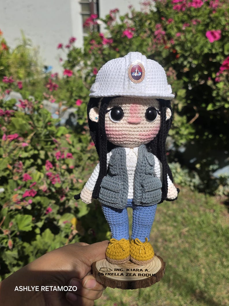 May include: A crocheted doll wearing a white hard hat with a circular emblem, a grey utility vest over a white shirt, blue pants, and yellow shoes. The doll stands on a circular wooden base engraved with the text ING. KIARA A. ESTRELLA ZEA ROQUE.