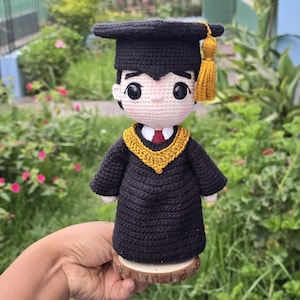 以下が含まれることがあります： 黒い帽子とガウンを着て、金色の縁取りが施された、かぎ針編みの卒業式のフィギュア。フィギュアは木製の台座の上に立っています。