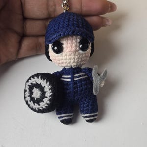 Może przedstawiać: Szydełkowy brelok do kluczy z lalką mechanika. Lalka ma na sobie niebieski mundur i czapkę, trzyma oponę i klucz. Lalka ma duże czarne oczy i jasnoróżową twarz. Brelok jest przymocowany do srebrnego metalowego kółka.