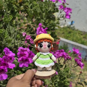 May include: A handmade crochet doll with red hair, wearing a green dress and yellow hat, stands on a small wooden slice. The doll is set against a backdrop of vibrant purple flowers and green foliage.