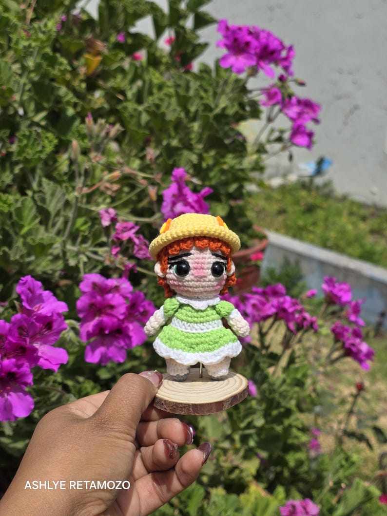 May include: A handmade crochet doll with red hair, wearing a green dress and yellow hat, stands on a small wooden slice. The doll is set against a backdrop of vibrant purple flowers and green foliage.