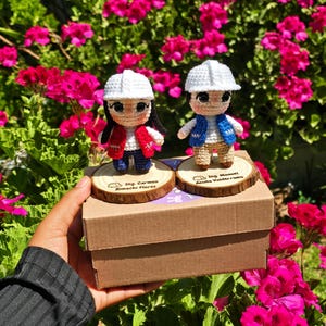 May include: Two crocheted dolls wearing white hard hats, standing on circular wooden bases atop a cardboard box. The left doll wears a red vest and blue pants, with text reading Ing. Carmen Rimachi Flores. The right doll wears a blue vest and tan pants, with text reading Ing. Manuel Acuña Valderrama.