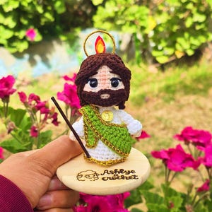 May include: A handmade crochet figure of a religious figure with brown hair and a beard, wearing a white robe and green sash. The figure is mounted on a small wooden base with the text "Ashlye crochet". A gold halo and staff are also included.