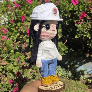 May include: A crochet doll with long black hair, wearing a white hard hat with a circular emblem, a white long-sleeved shirt, blue pants, and yellow shoes. The doll stands on a circular wooden base with the text ING. KIARA A. ESTRELLA ZEA ROQUE.