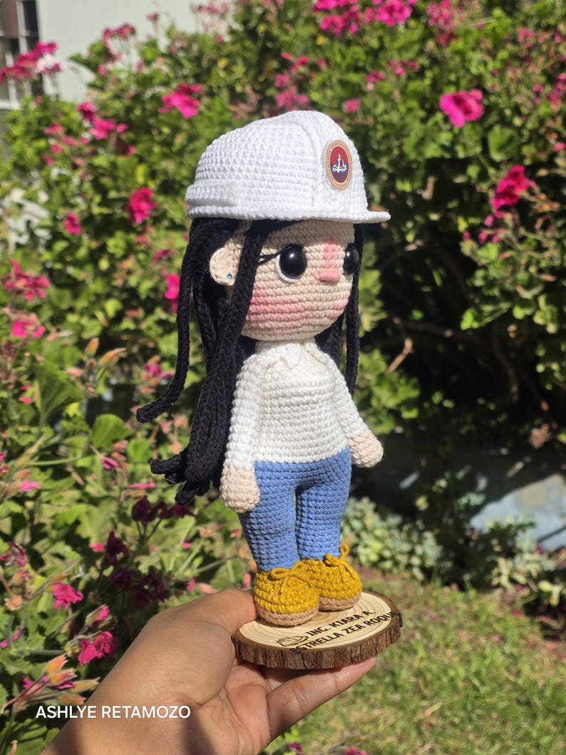 May include: A crochet doll with long black hair, wearing a white hard hat with a circular emblem, a white long-sleeved shirt, blue pants, and yellow shoes. The doll stands on a circular wooden base with the text ING. KIARA A. ESTRELLA ZEA ROQUE.