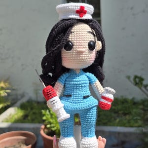 Pu&ograve; includere: Una bambola all'uncinetto fatta a mano che raffigura un infermiere con lunghi capelli neri, che indossa un top e pantaloni da infermiere azzurri, scarpe bianche e una cuffia da infermiere bianca con una croce rossa.