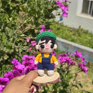 May include: A handmade crochet doll of a person wearing a yellow shirt, blue overalls, and a green hat. The doll is standing on a small wooden slice, with a background of green foliage and purple flowers.