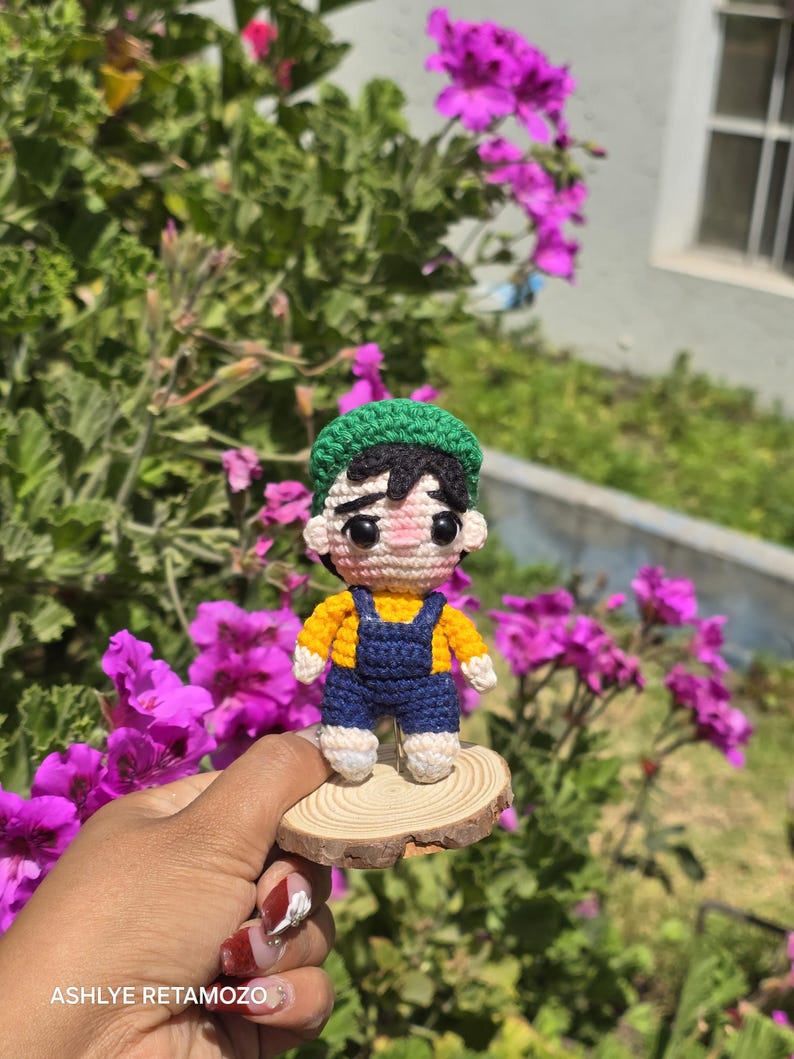 May include: A handmade crochet doll of a person wearing a yellow shirt, blue overalls, and a green hat. The doll is standing on a small wooden slice, with a background of green foliage and purple flowers.