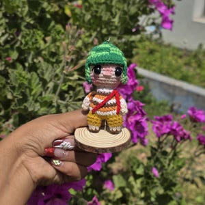 May include: Crocheted doll of a cartoon character wearing a green hat, striped shirt, and yellow pants. The doll is mounted on a small wooden disc. The background features green foliage and purple flowers.