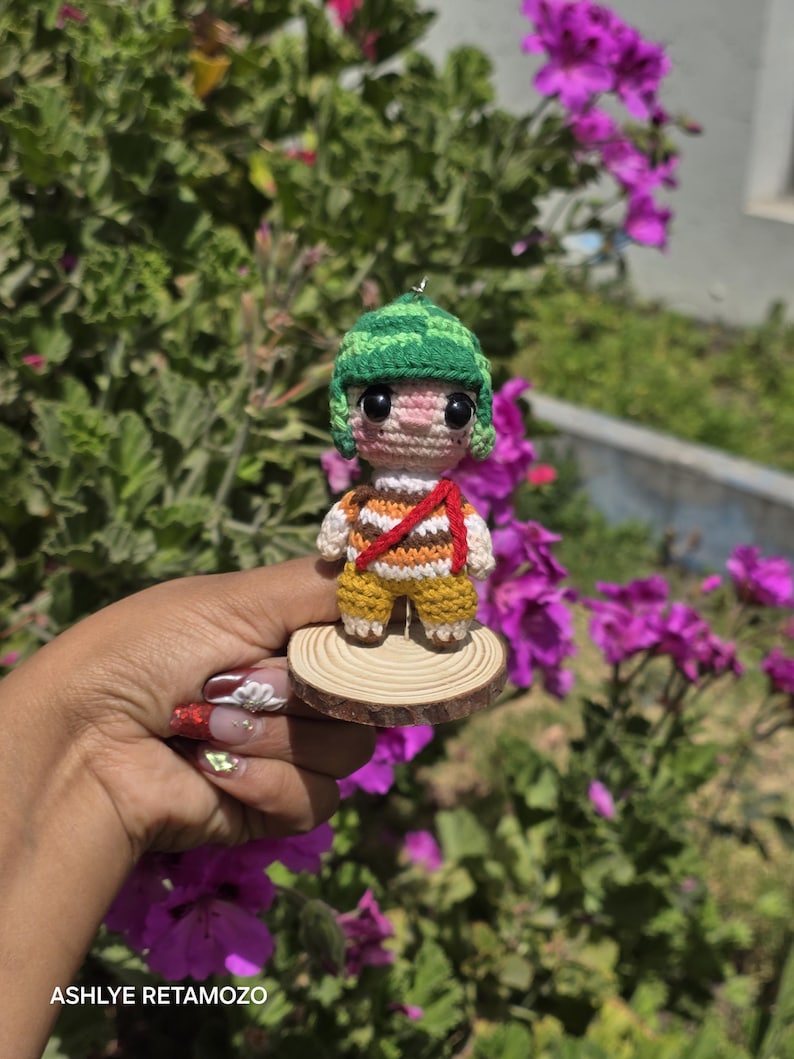 May include: Crocheted doll of a cartoon character wearing a green hat, striped shirt, and yellow pants. The doll is mounted on a small wooden disc. The background features green foliage and purple flowers.