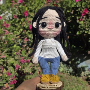 May include: A handmade crochet doll with long black yarn hair, wearing a white long-sleeved shirt, blue pants, and yellow shoes. The doll stands on a circular wooden base engraved with the text ING. KIARA A. ESTRELLA ZEA ROQUE.