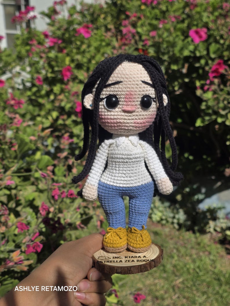 May include: A handmade crochet doll with long black yarn hair, wearing a white long-sleeved shirt, blue pants, and yellow shoes. The doll stands on a circular wooden base engraved with the text ING. KIARA A. ESTRELLA ZEA ROQUE.