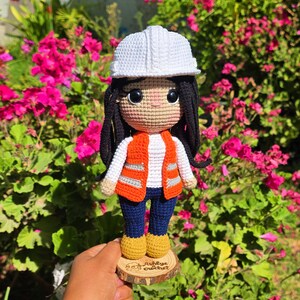 May include: A crocheted doll featuring a white hard hat, long black hair, a white long-sleeved shirt, an orange high-visibility vest with grey reflective stripes, dark blue pants, and yellow boots, standing on a wooden base labeled Ashlye crochet.