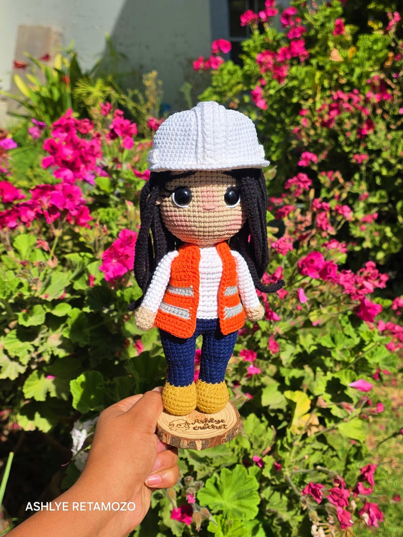 May include: A crocheted doll featuring a white hard hat, long black hair, a white long-sleeved shirt, an orange high-visibility vest with grey reflective stripes, dark blue pants, and yellow boots, standing on a wooden base labeled Ashlye crochet.