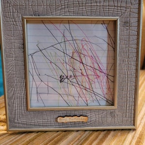 May include: A gray picture frame with a white mat and a drawing inside. The drawing is a child's scribble with colorful lines and the word "Ben" written in black marker. The frame has a wooden plaque on the bottom that says "Benji DaMonkey".