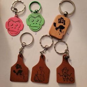 May include: Five different keychains with various designs. Keychain 1 is pink with a monkey face. Keychain 2 is green with a monkey face. Keychain 3 is wooden with a handprint. Keychain 4 is brown leather with a handprint. Keychain 5 is brown leather with a heart and two monkeys. Keychain 6 is brown leather with a monkey.