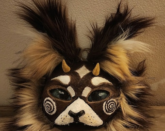 May include: A brown and white furry mask with a cat-like face, featuring large eyes, a small nose, and two small horns. The mask has a swirling pattern around the eyes.