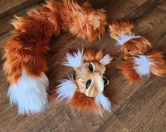 May include: A cat costume set featuring a mask, tail, and paw covers. The mask is tan with green eyes and a black mesh mouth. The tail and paw covers are orange and white, with a fluffy texture. The set is designed for cosplay or costume use.