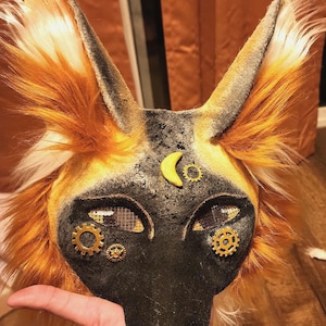 May include: A black and yellow fox mask with a steampunk design. The mask has a crescent moon and gears on the forehead, and mesh eyes with gears around them.