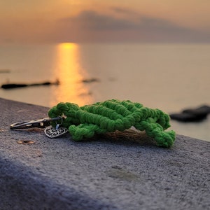 May include: A green macrame turtle keychain with a silver-colored heart charm. The keychain rests on a gray surface with a sunset over the ocean in the background.