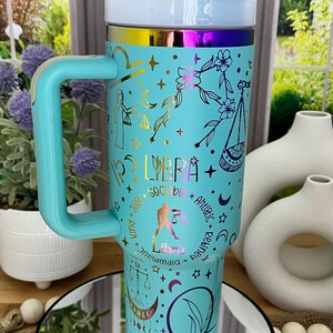 May include: A turquoise travel mug with a handle, featuring Libra zodiac symbols and text. The mug has a white lid and a rainbow-colored band. The design includes stars, moons, and floral elements. The text includes "Libra", "Sociable", and "Refined".